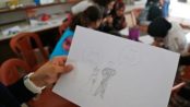 A child holds up a drawing in a Syrian refugee camp where she shows a young bride appearing sad and thinking of school, while a groom smiles and thinks of a home. Creative commons image.